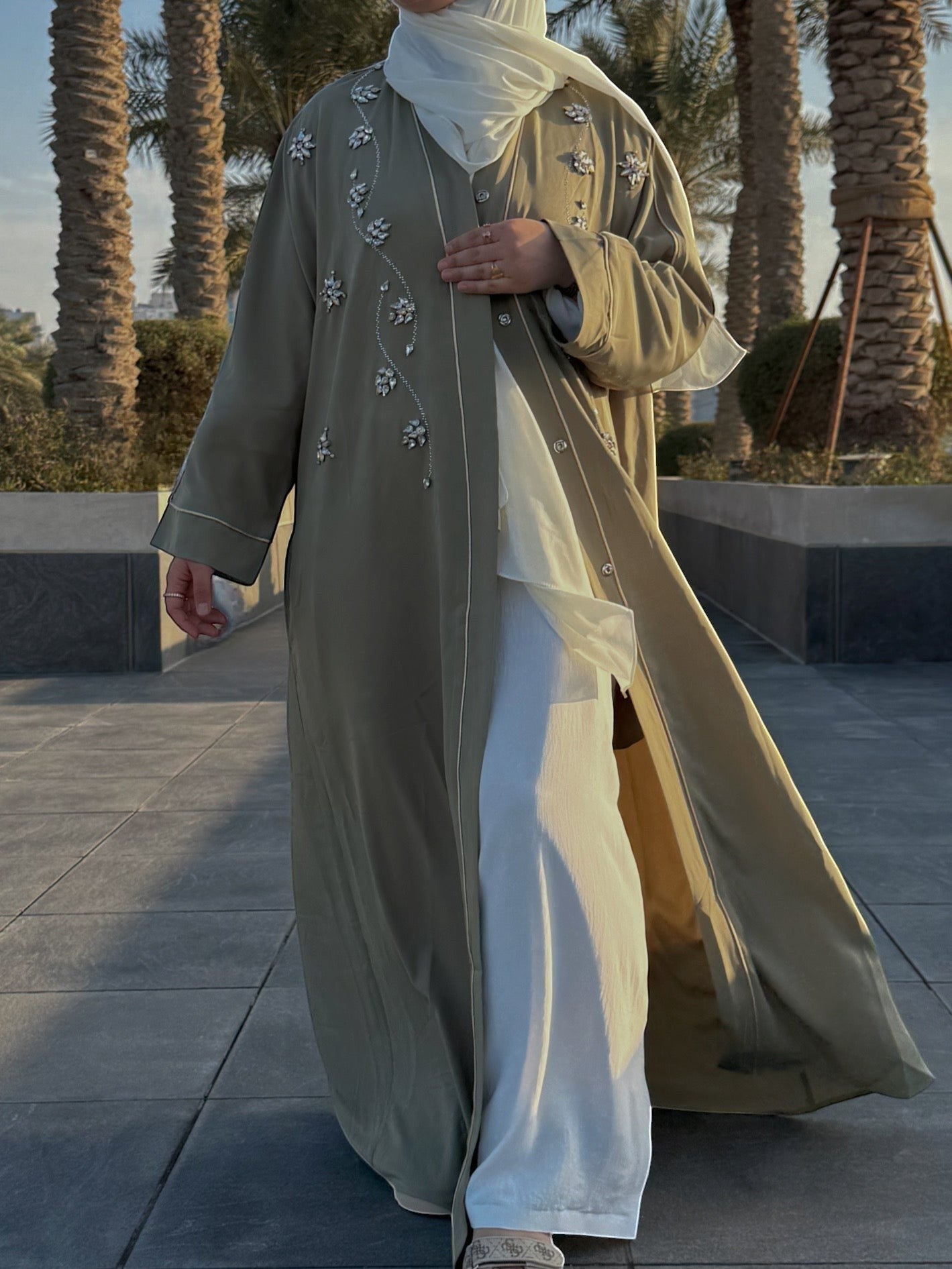 Nesthetics Prestige Abaya in olive green with white trim and floral crystal embellishments. Worn with white trousers and hijab.